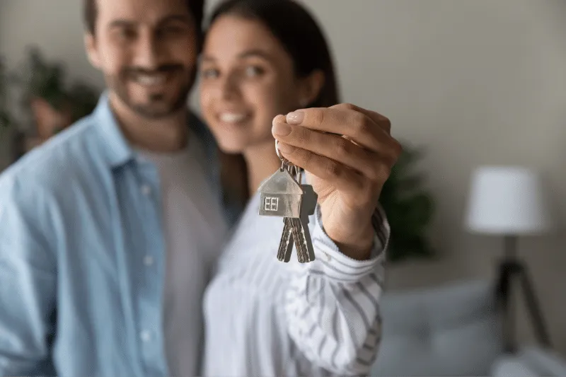 A smiling couple stands close together, with the woman holding out a set of house keys toward the camera, suggesting they have just bought a new home. The background is softly blurred indoors.